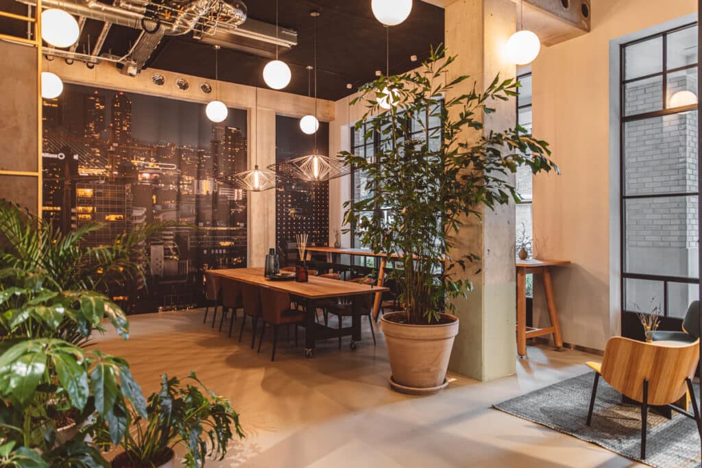 Lounge en overlegruimte in Rotterdam Little C met houten tafel, planten, hanglampen en stadsfoto aan de wand.