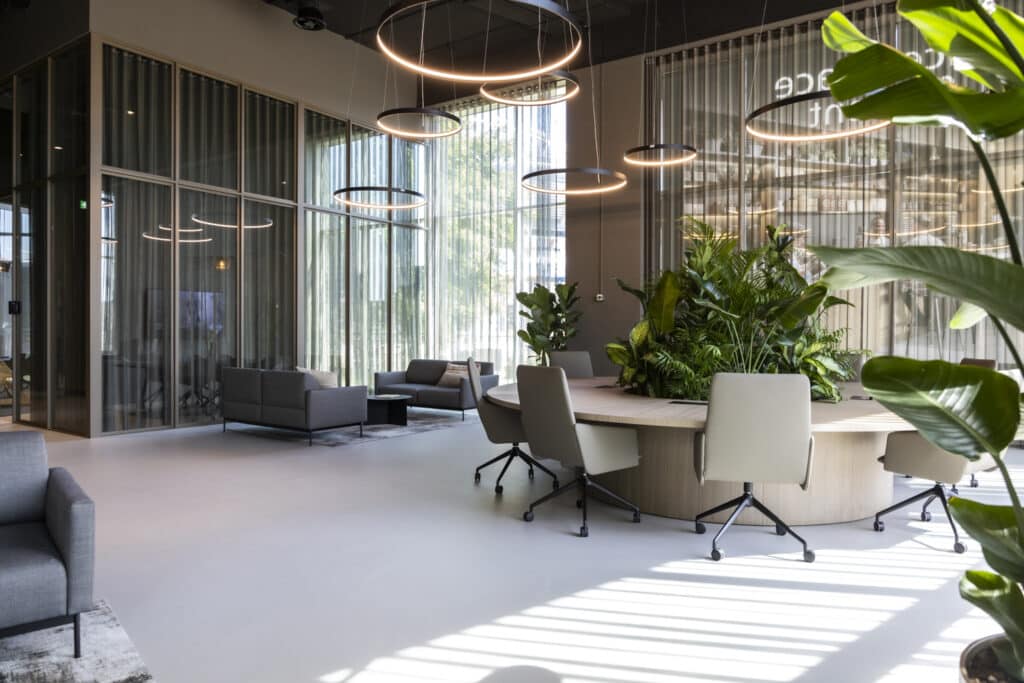Moderne lounge in Amsterdam Arena met ronde overlegtafel, grijze banken, planten en ringlampen.